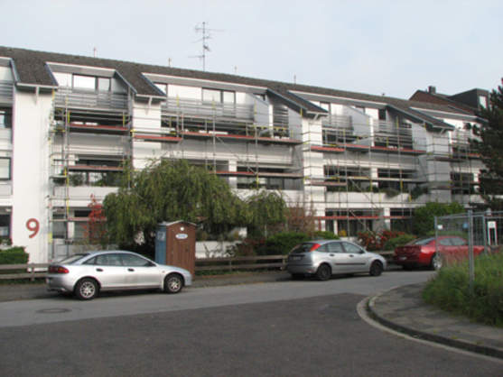 Mehrfamilienhaus mit Gerüst für Fassadensanierung, Autos parken vor dem Gebäude.